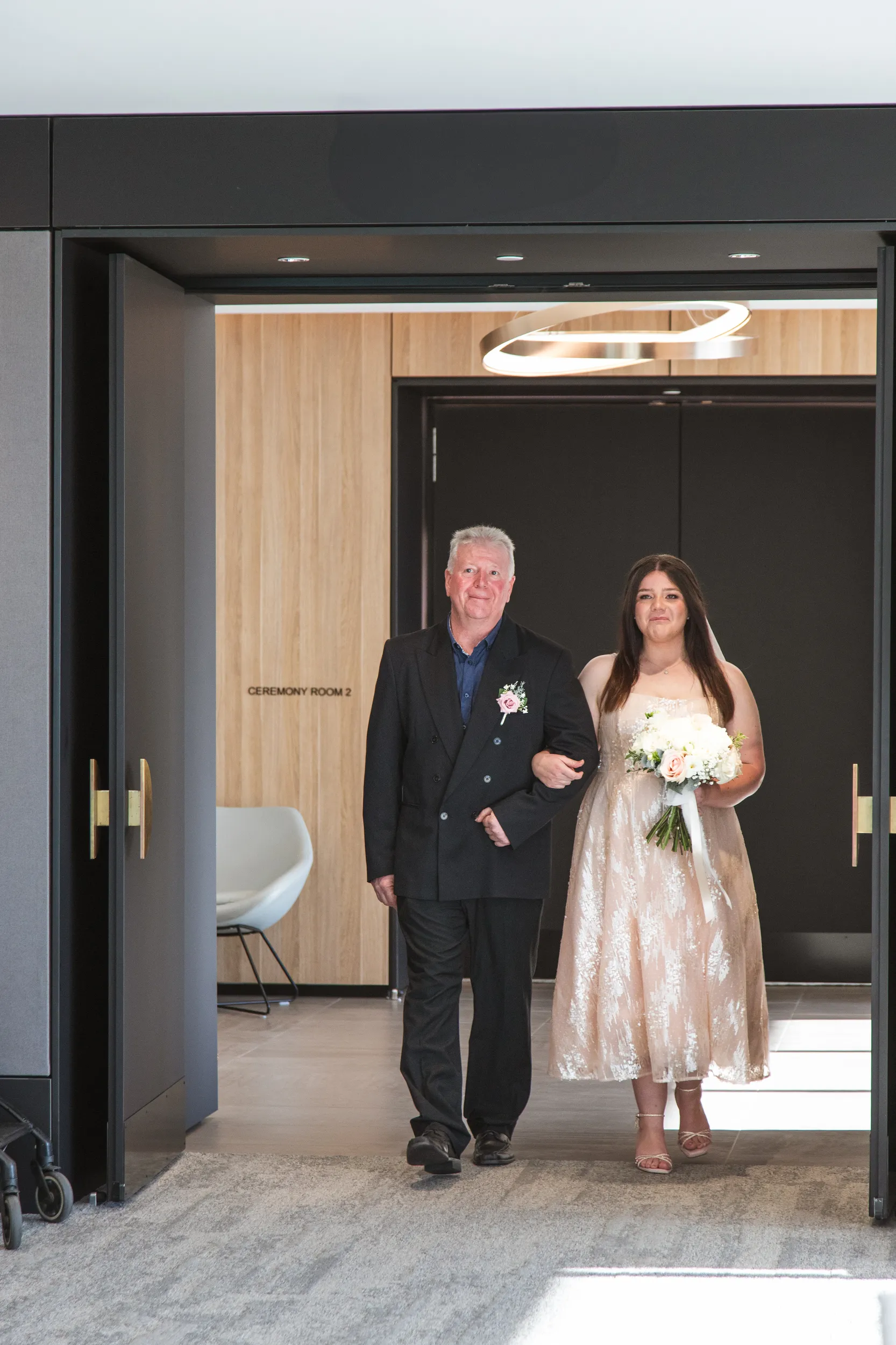 Carlie walking arm-in-arm with father outside Ceremony Room 2 in modern venue with contemporary architecture