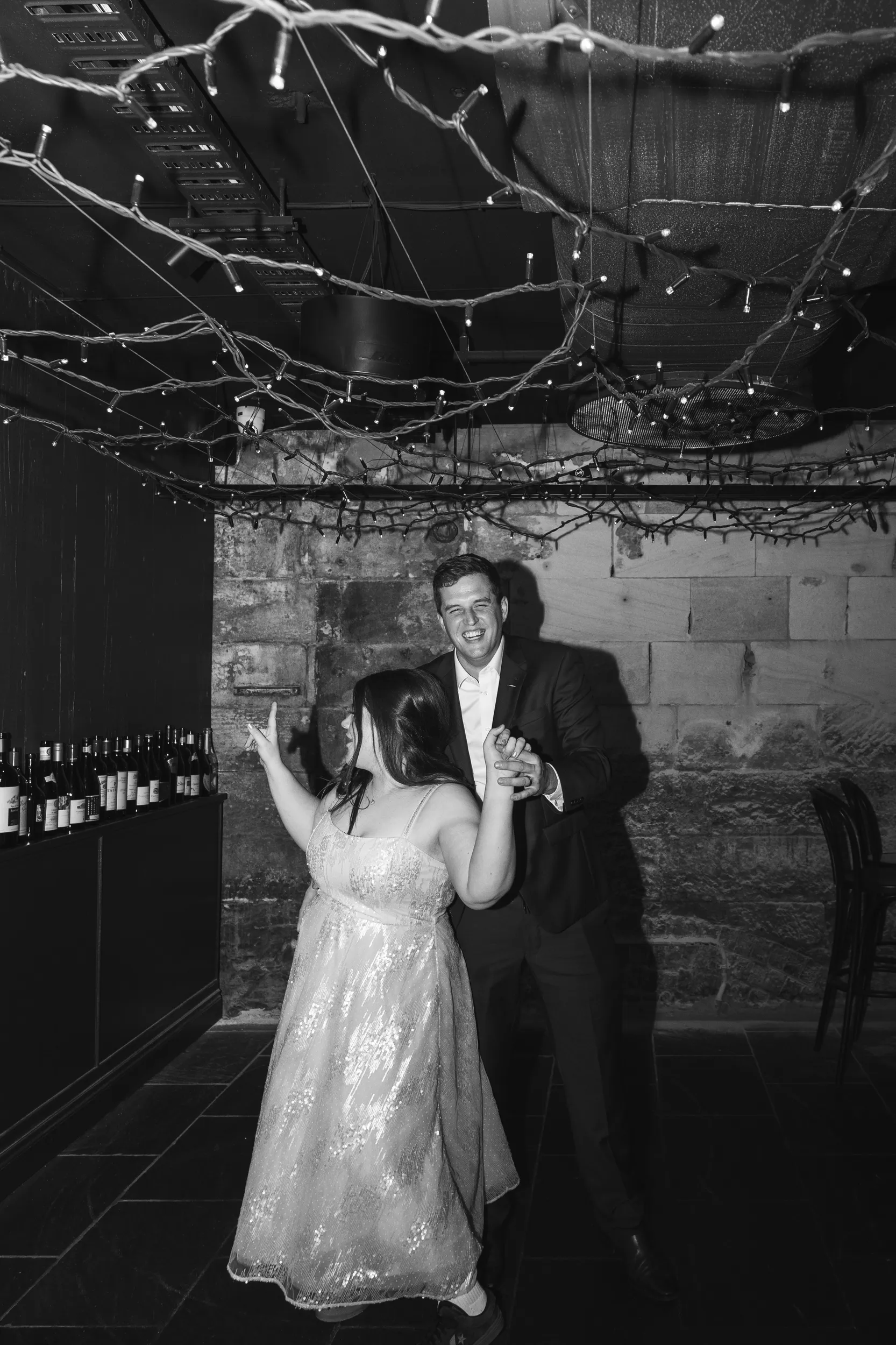 Carlie and Iain black and white dancing moment in rustic cellar with string lights and wine bottles