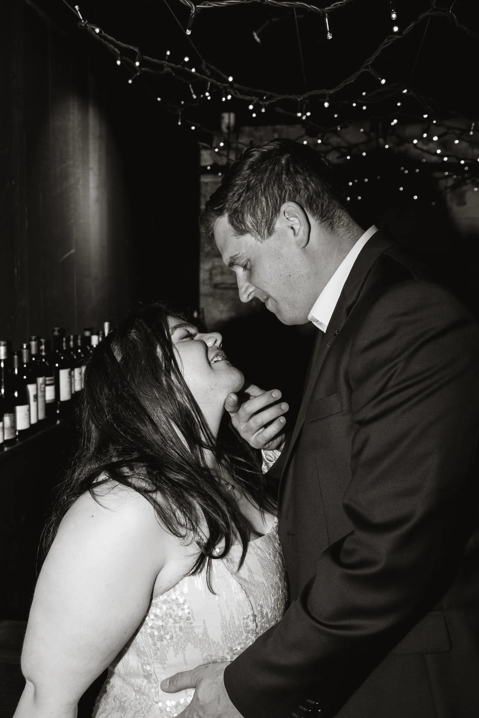 Carlie and Iain romantic black and white embrace in rustic cellar with string lights and wine bottles backdrop
