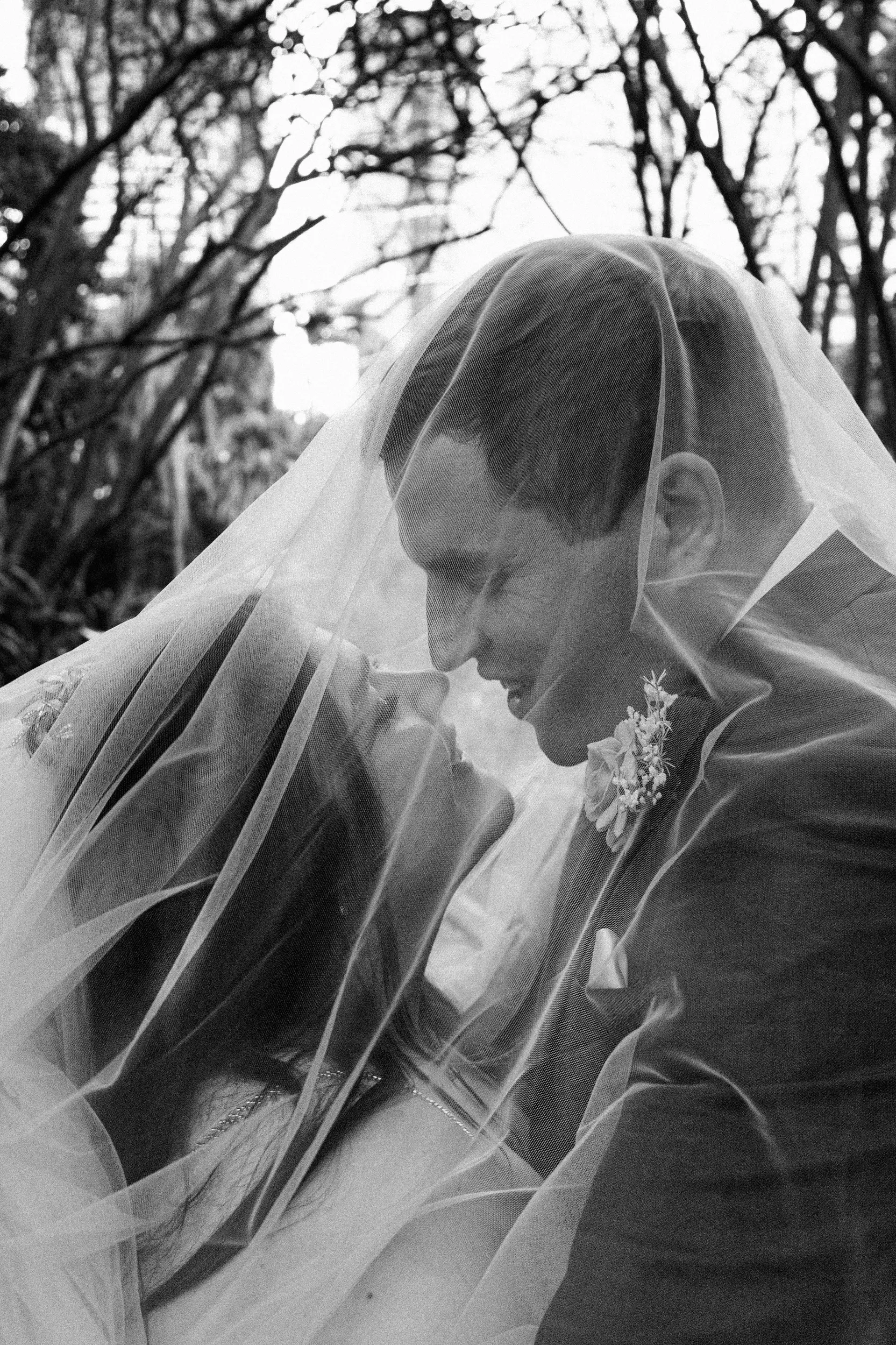 Carlie and Iain intimate black and white embrace with flowing veil under natural tree lighting