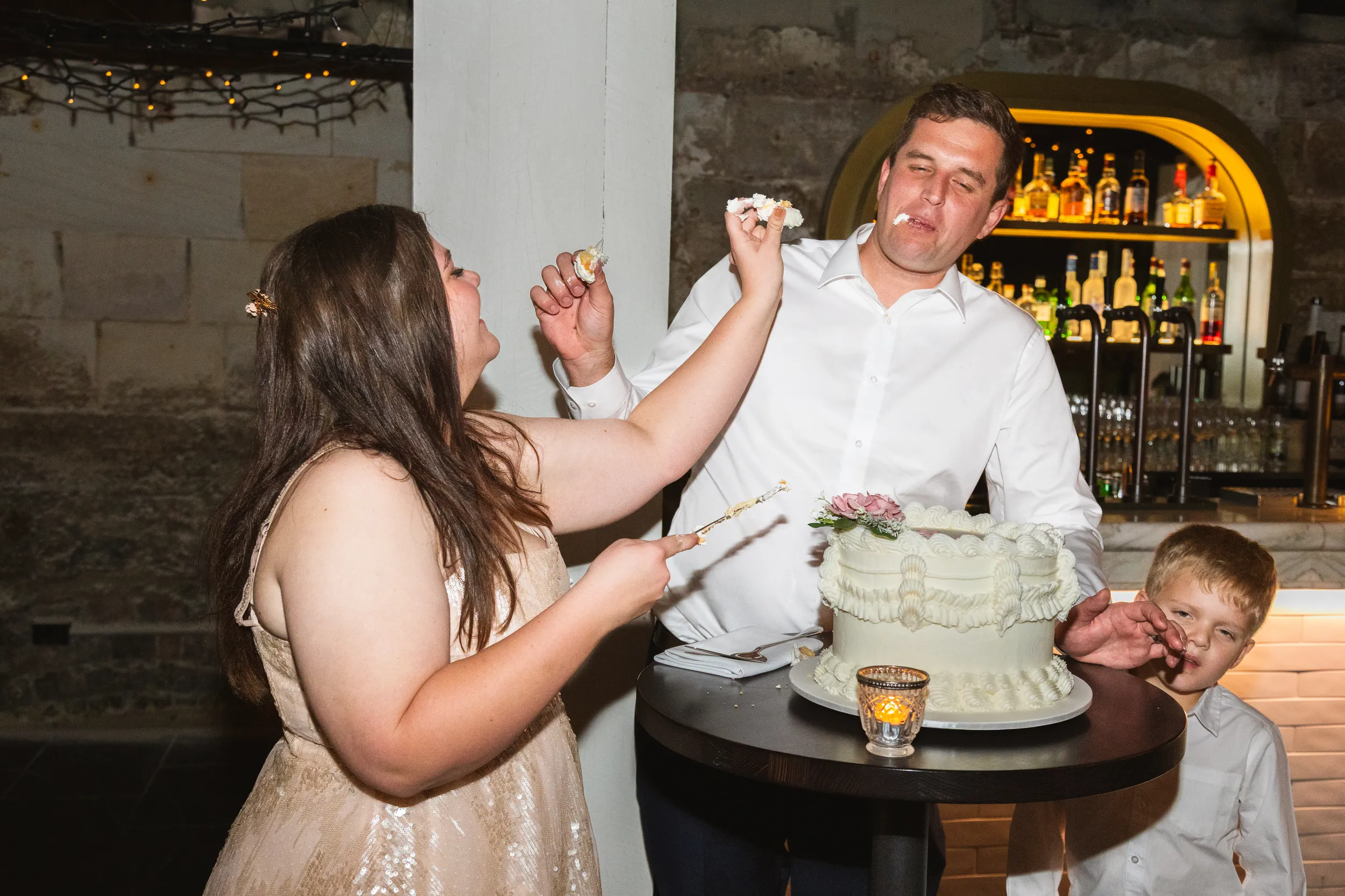 Carlie and Iain playful cake feeding moment in rustic venue with exposed brick walls and young child watching