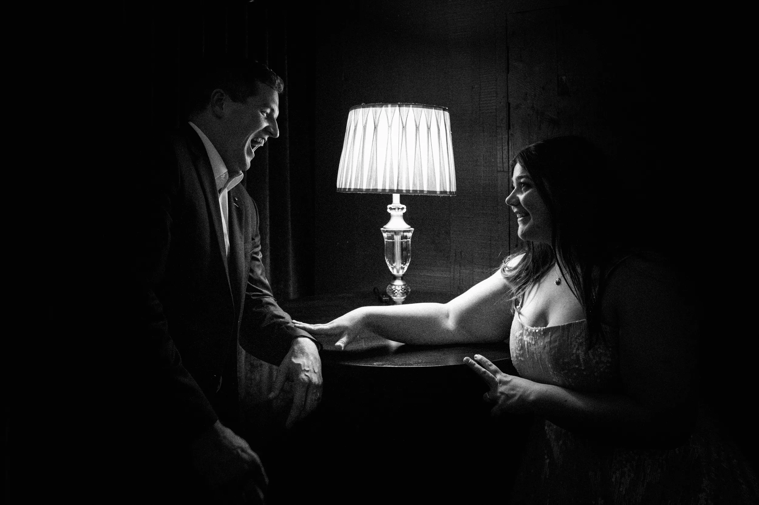 Carlie and Iain intimate black and white moment holding hands across table with vintage lamp providing dramatic lighting