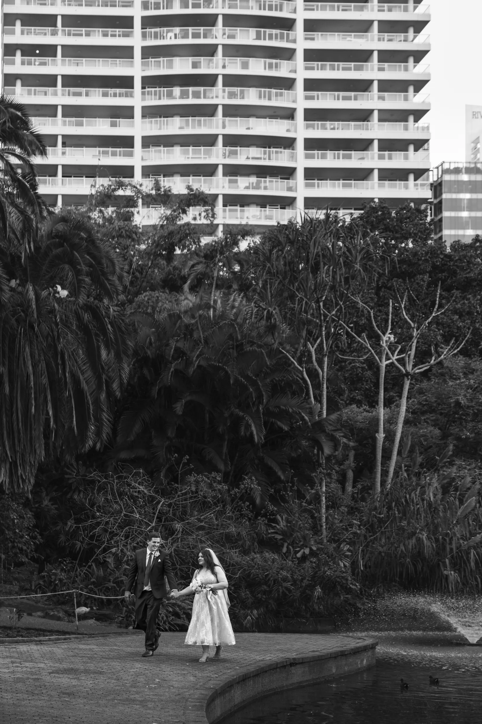 Carlie and Iain black and white urban portrait walking hand-in-hand with modern high-rise buildings and tropical landscaping