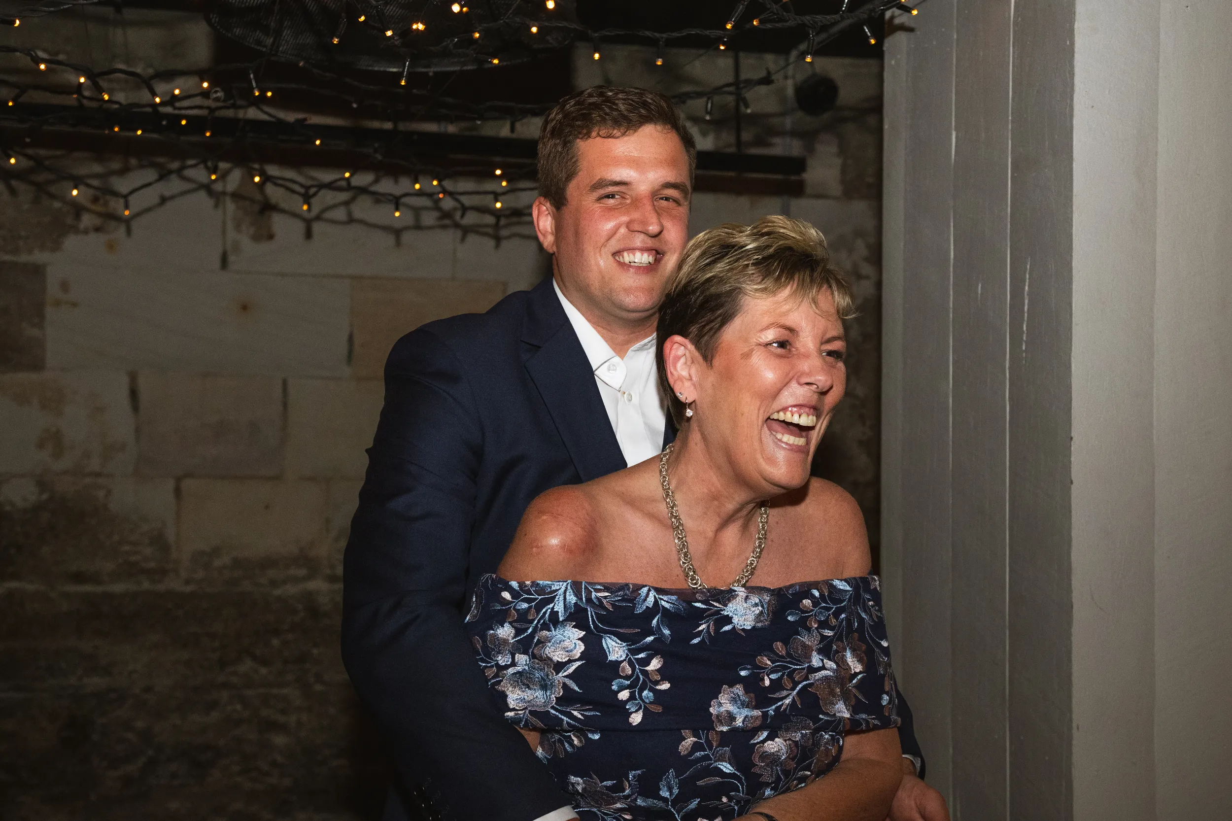 Iain joyful embrace with mother in rustic cellar setting with string lights during wedding celebration