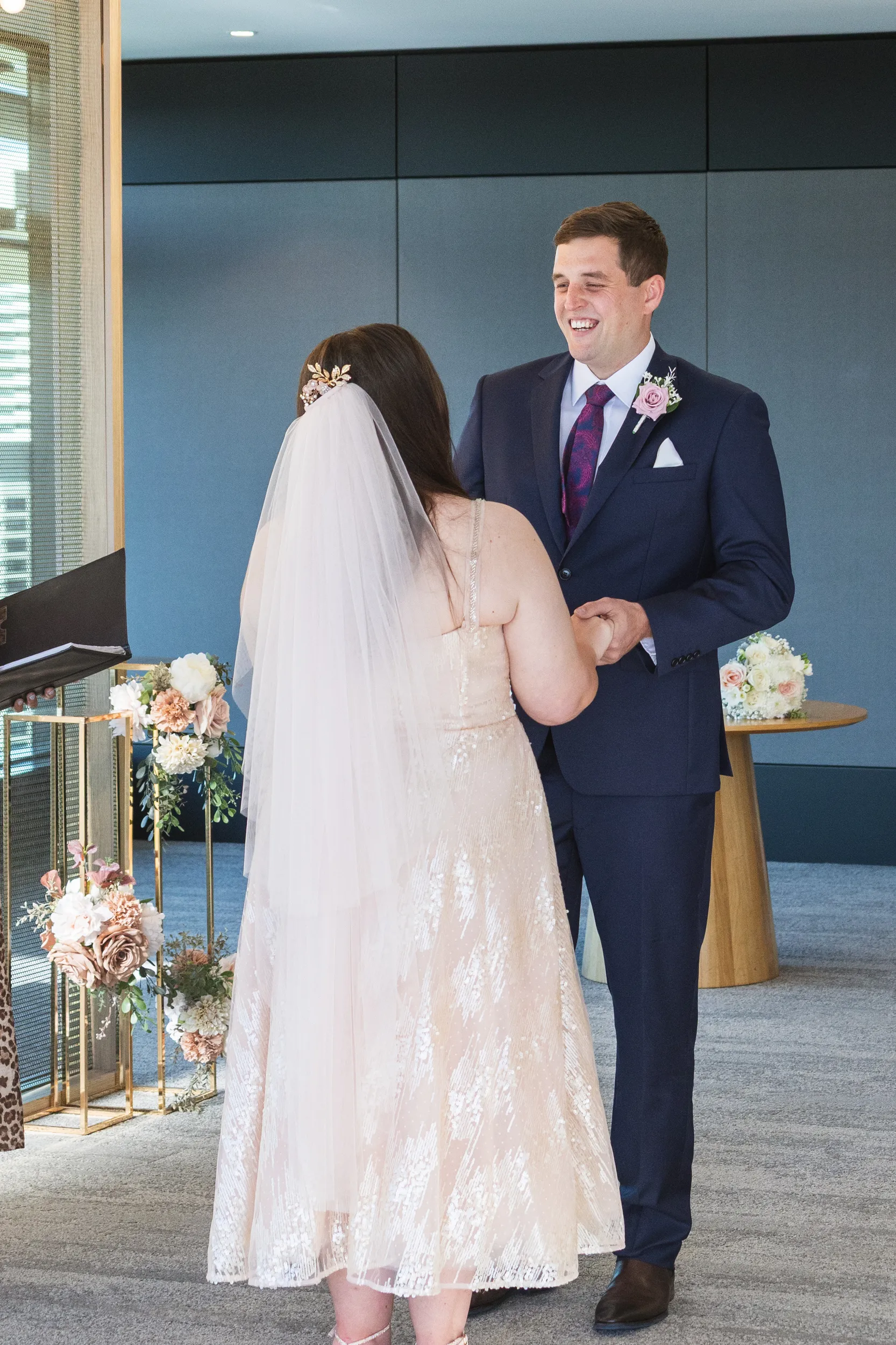 Iain joyful ceremony moment smiling while holding Carlie's hands in modern venue with floral arrangements
