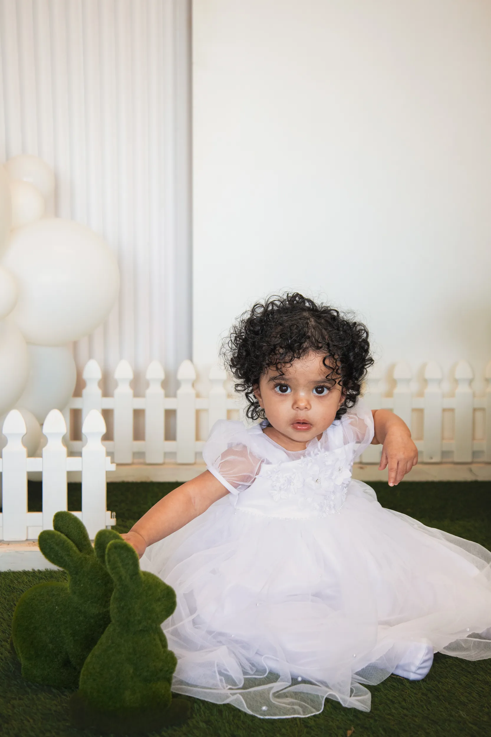 Adorable baptism portrait of child with curly hair in white tulle dress with picket fence backdrop