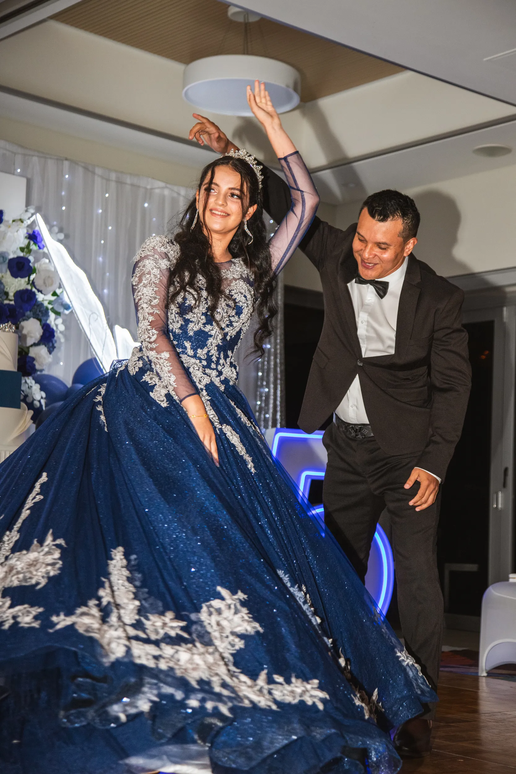 Joyful debut celebration moment with young woman in blue ball gown and tiara waving with both hands raised