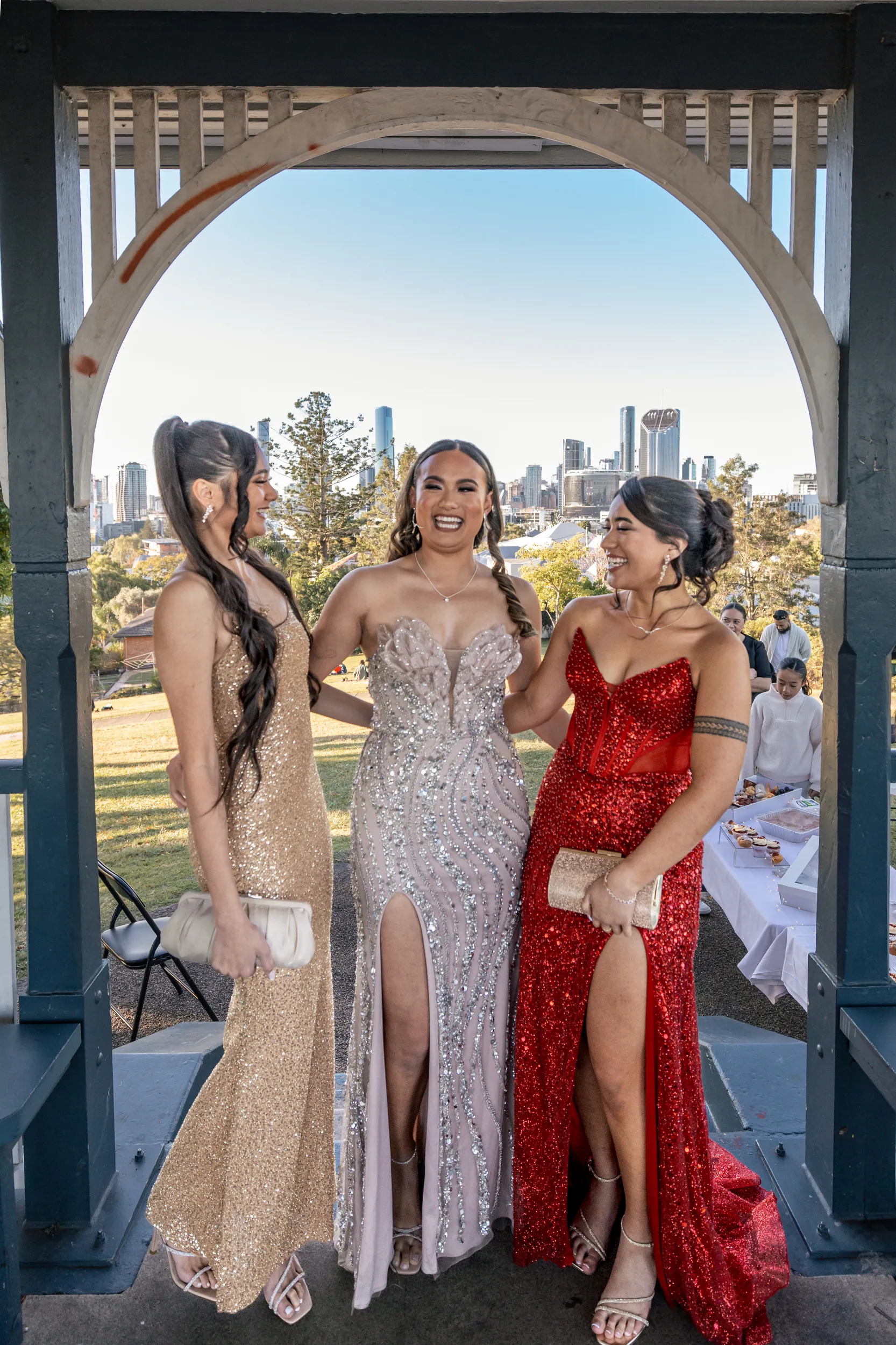Stunning formal group photo of three women in elegant gowns under architectural archway with city skyline