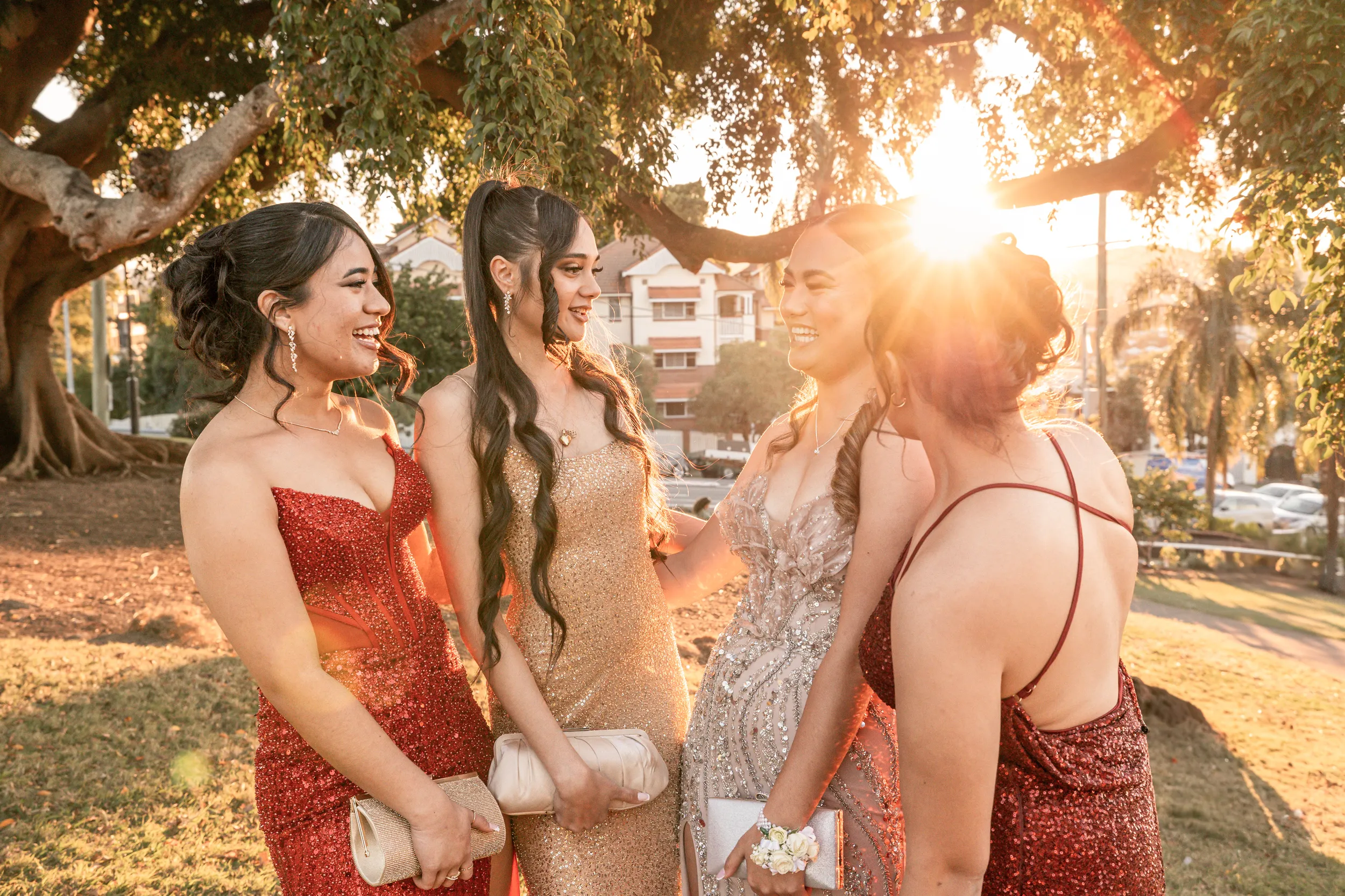 Joyful group photo of four women in elegant evening gowns during golden hour under large tree