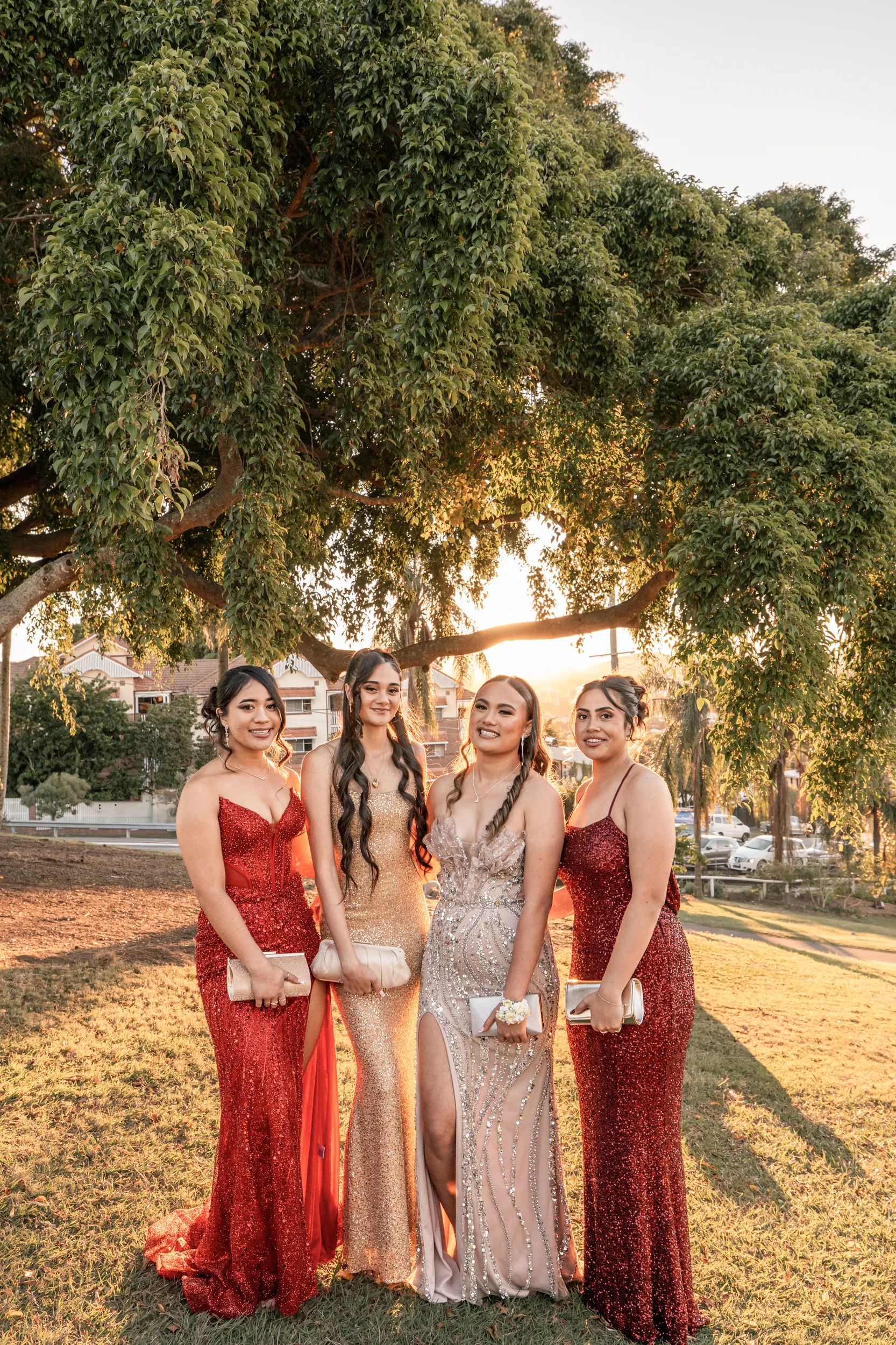 Gorgeous formal group photo of four women in elegant gowns under large tree during golden hour