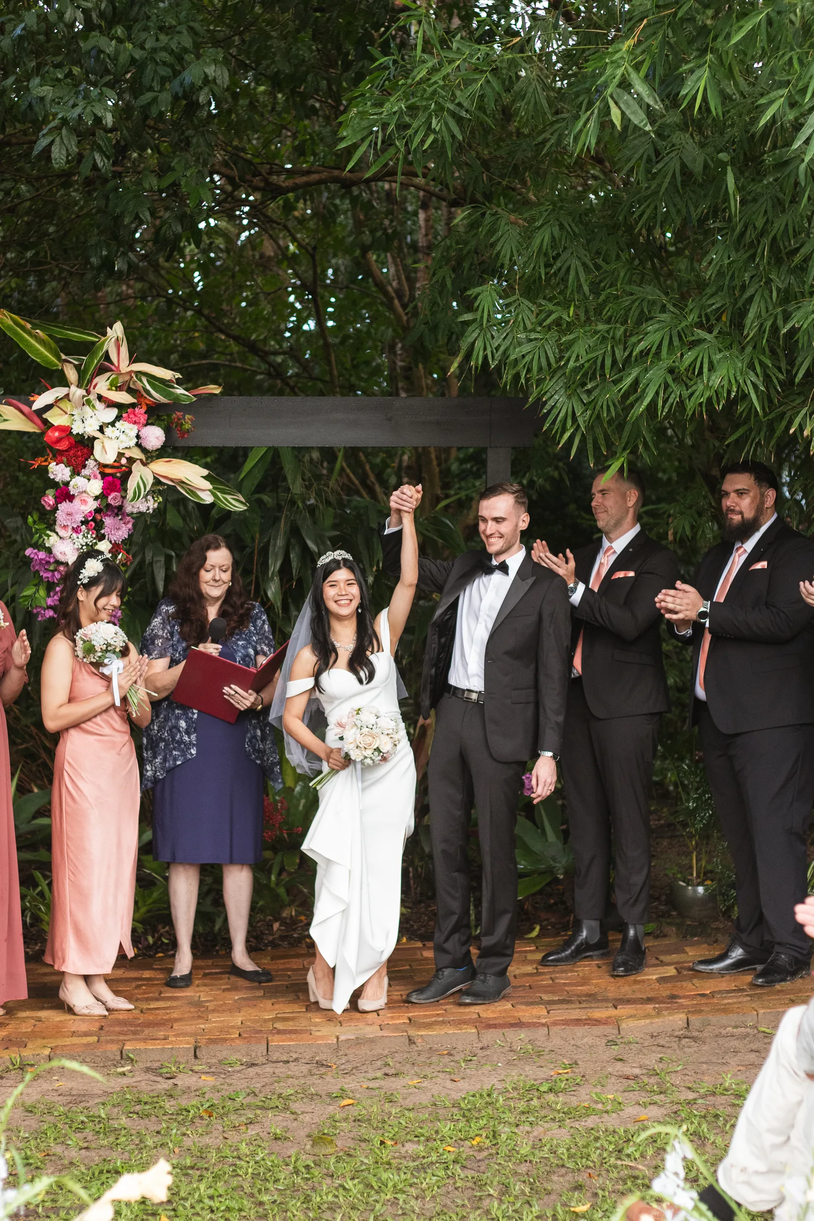 Triumphant moment after wedding ceremony with couple raising hands in celebration under floral arch with wedding party applauding