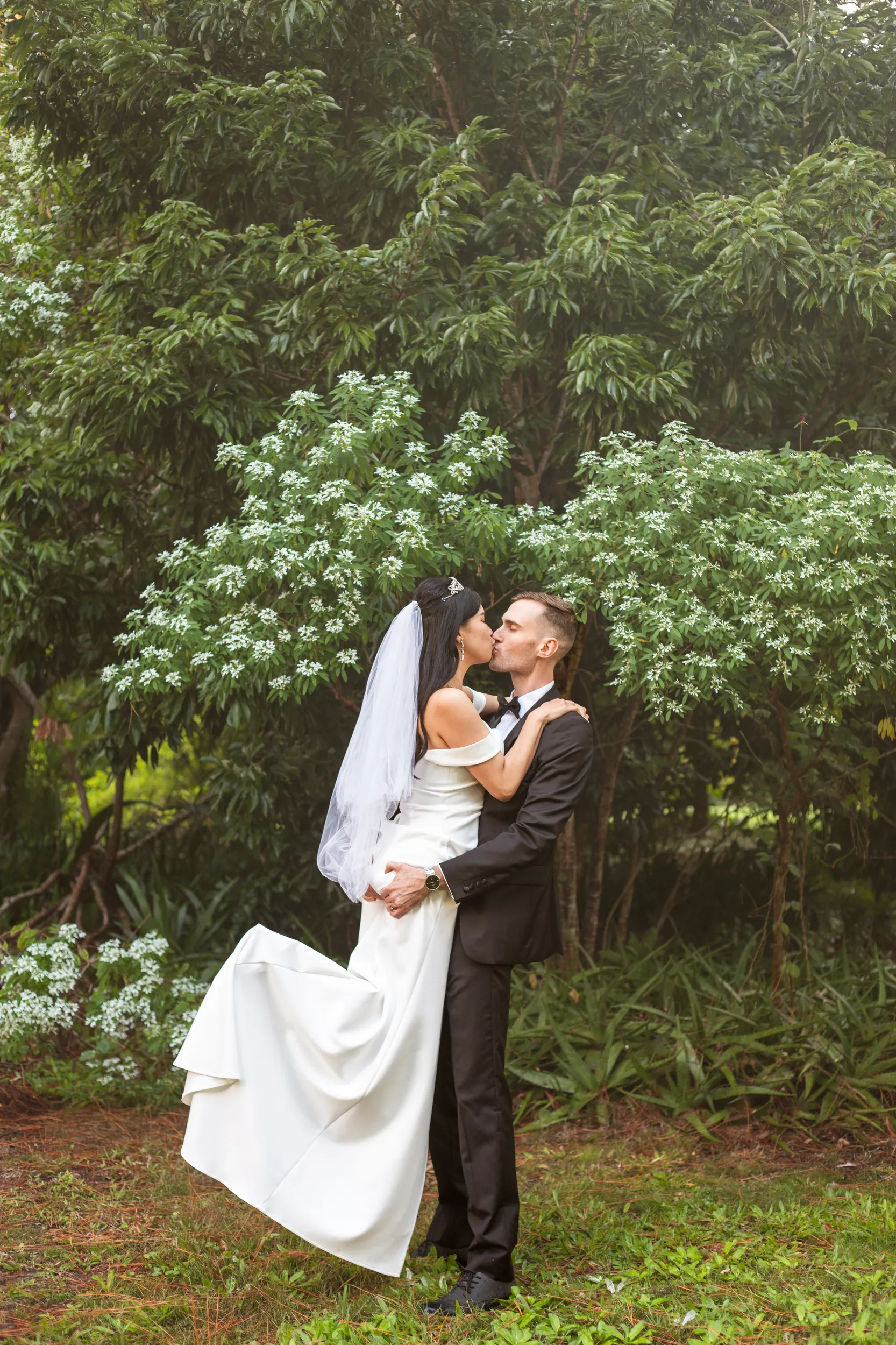 Huiyu and Chris romantic kiss in garden with bride in white dress and veil surrounded by lush greenery and white flowers