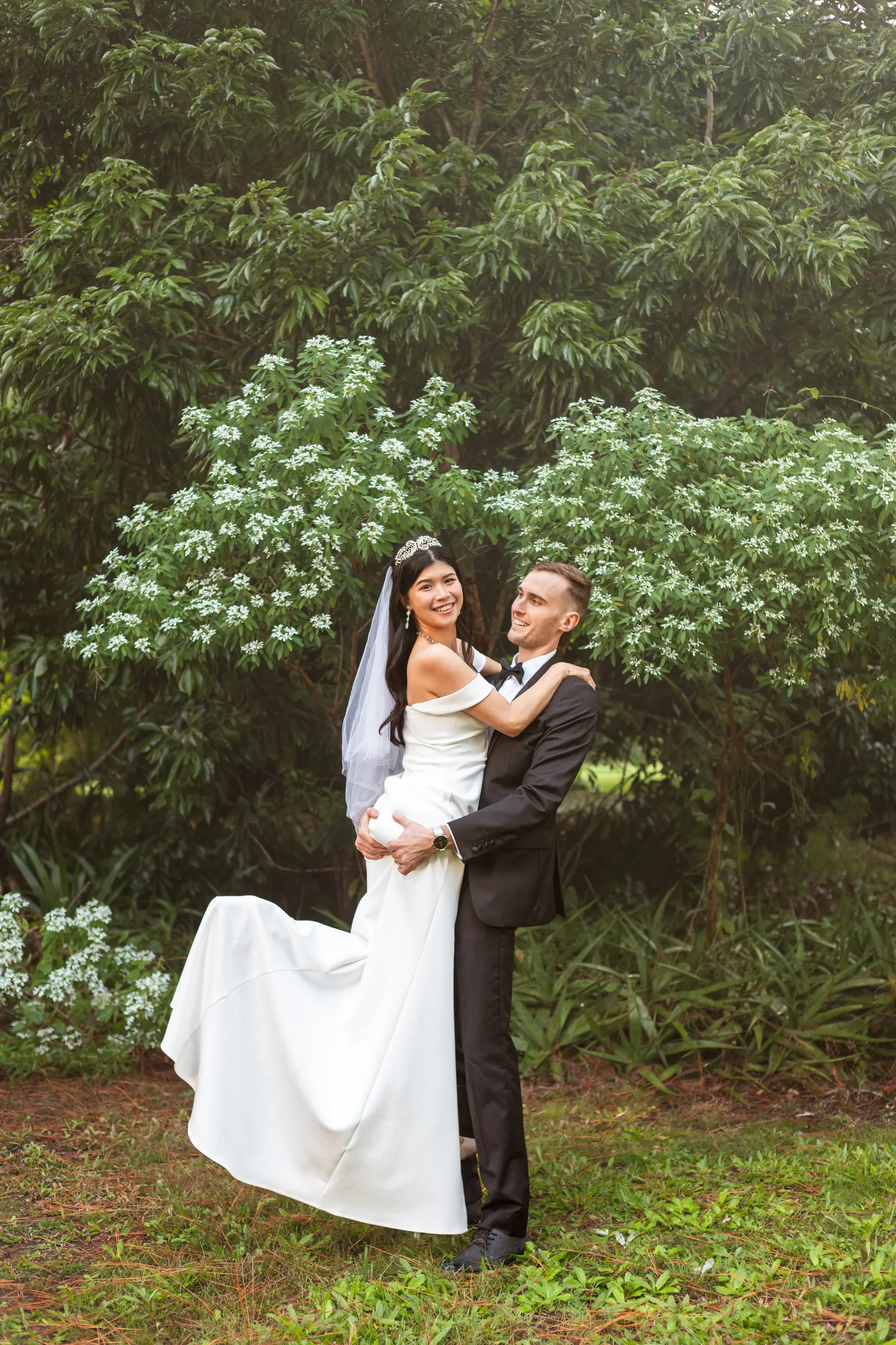 Huiyu and Chris romantic portrait with groom lifting bride against lush green backdrop with white flowering bushes