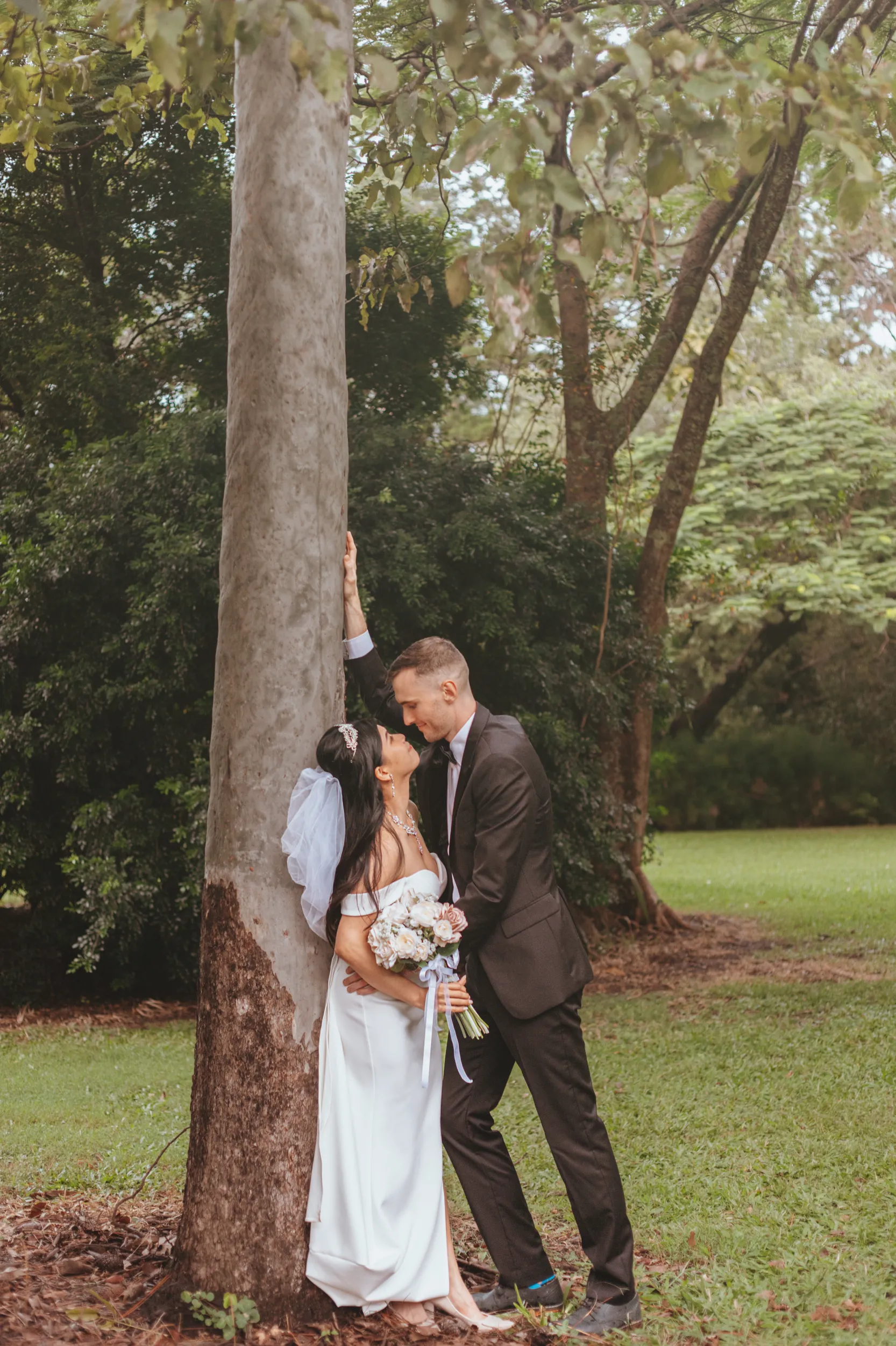Huiyu and Chris romantic portrait against large tree trunk with groom's hand on tree in natural garden setting