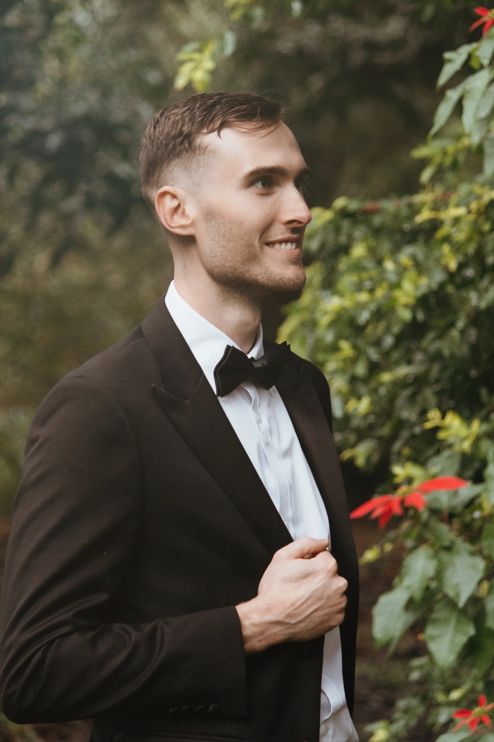 Chris handsome groom portrait in black tuxedo with bow tie smiling in garden setting with red flowers