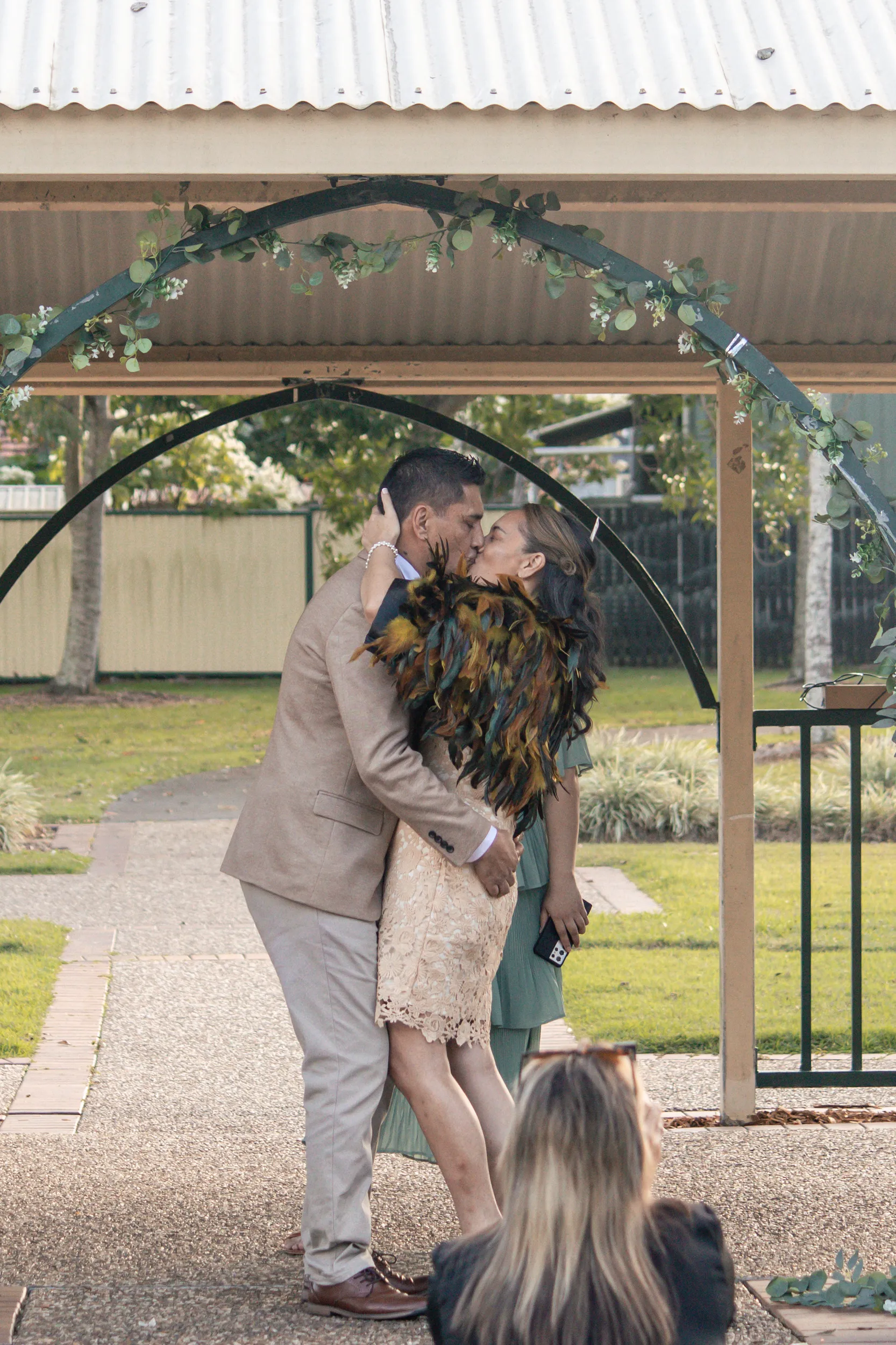 Romantic wedding kiss under pavilion floral arch with traditional feather cloak and guests