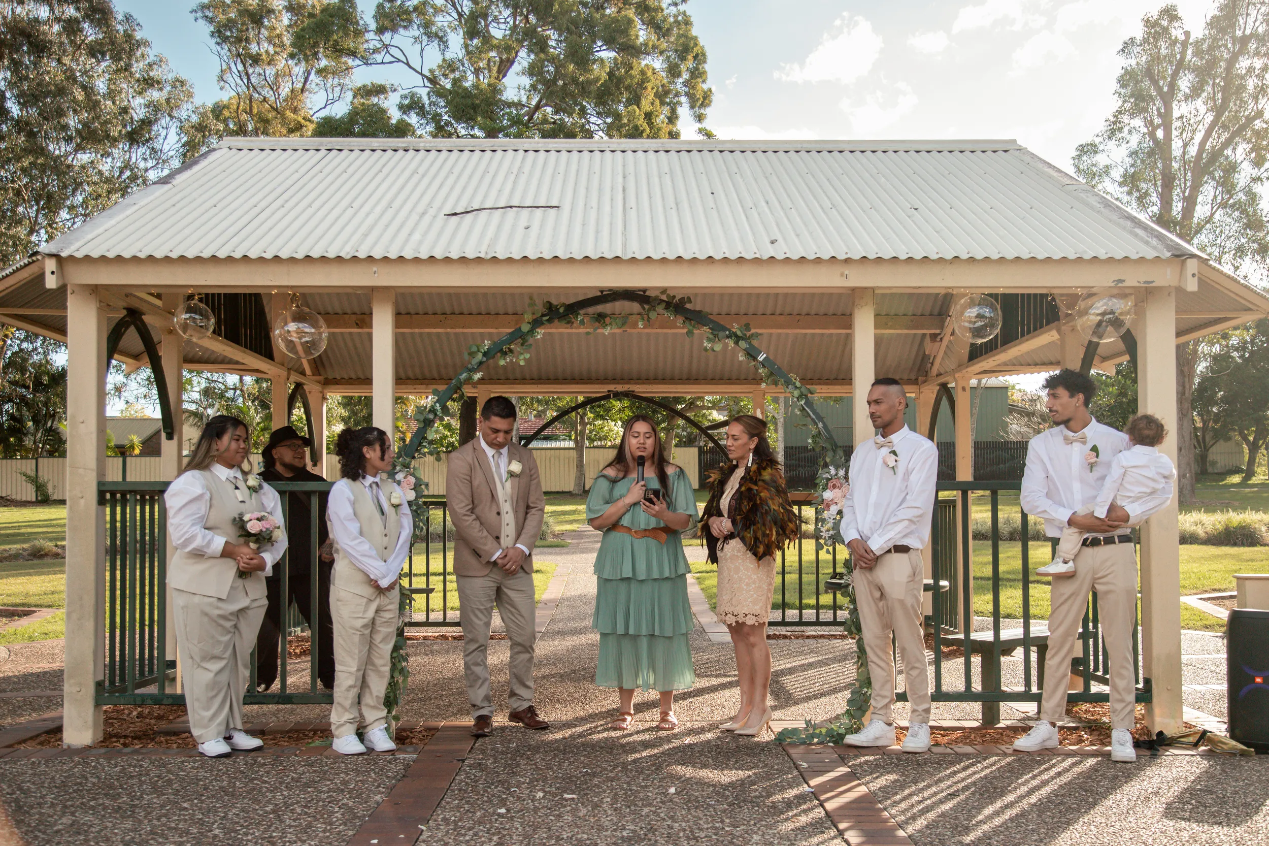 Outdoor wedding ceremony under rustic pavilion with floral arch and wedding party in neutral tones