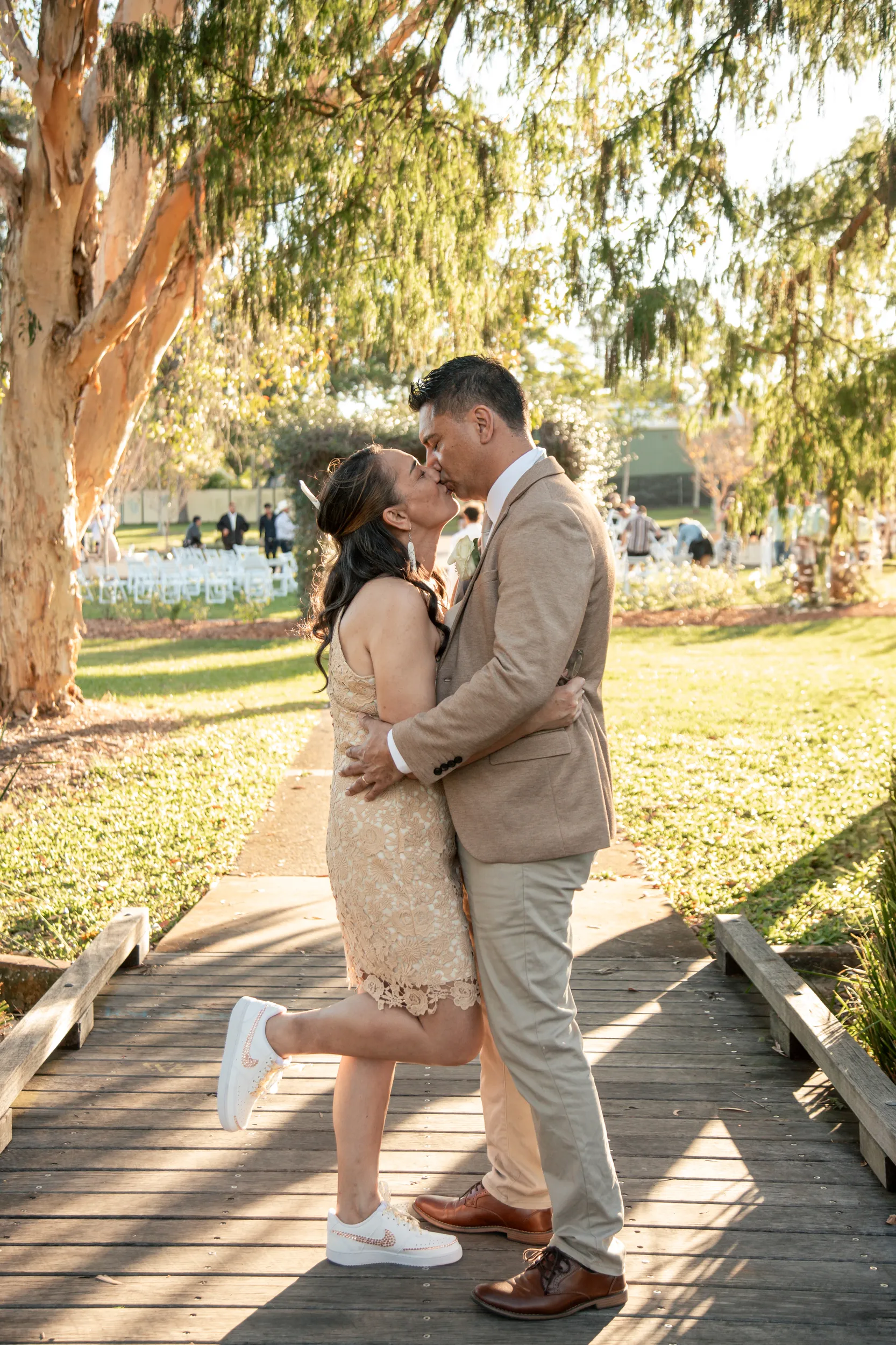 Romantic wedding moment with couple kissing on wooden boardwalk under weeping willow trees during golden hour