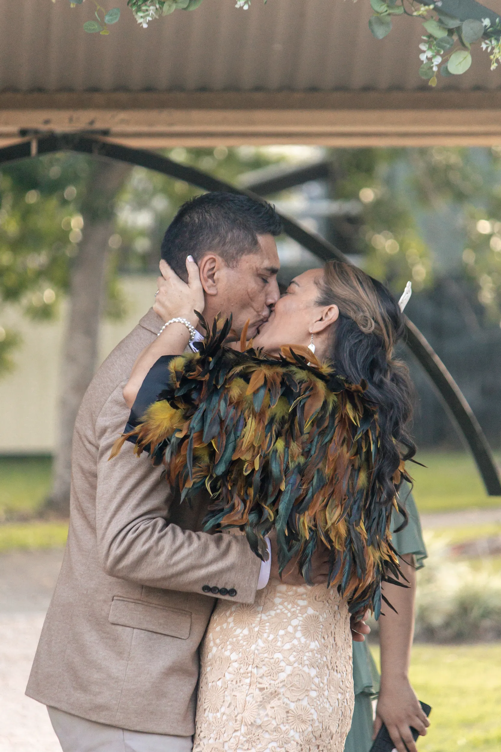 Intimate wedding kiss under floral arch with bride in traditional Māori feather cloak and groom in beige suit