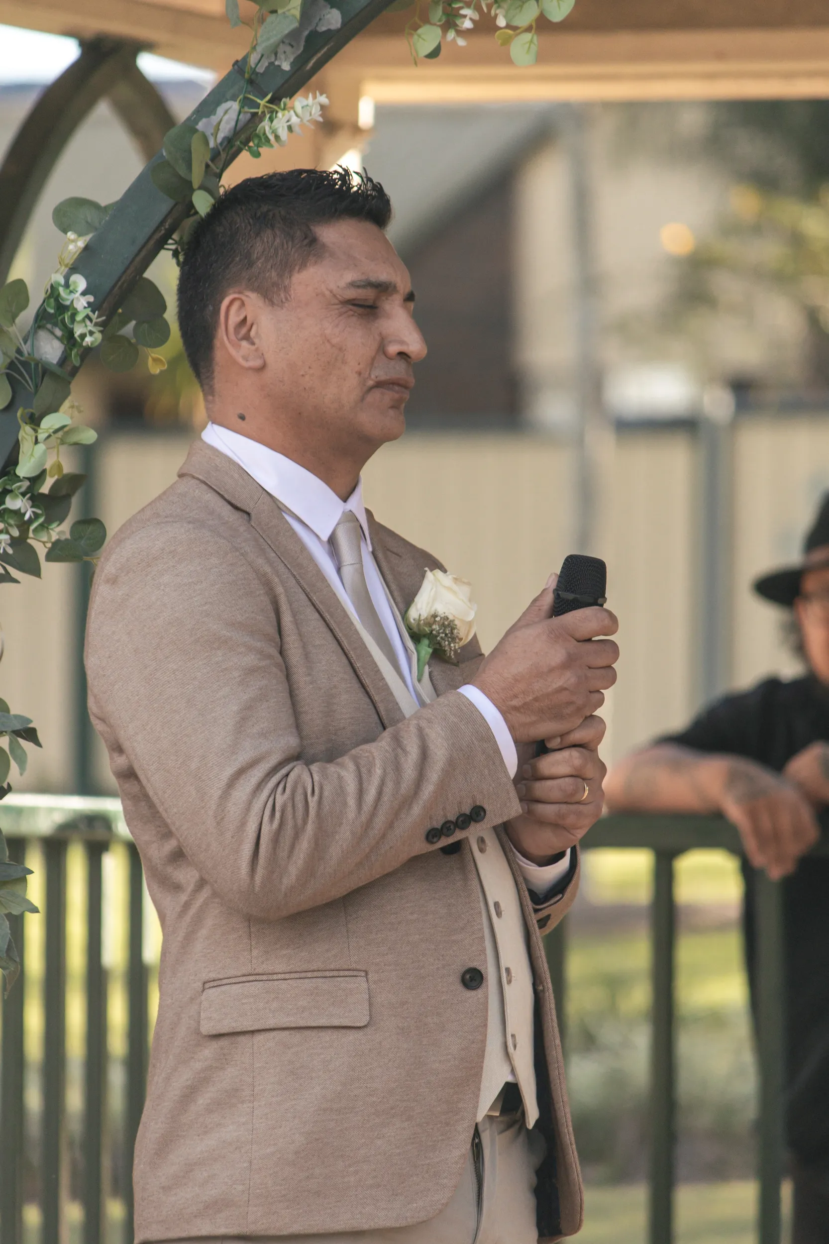 Groom giving heartfelt wedding speech with microphone under floral arch in beige three-piece suit