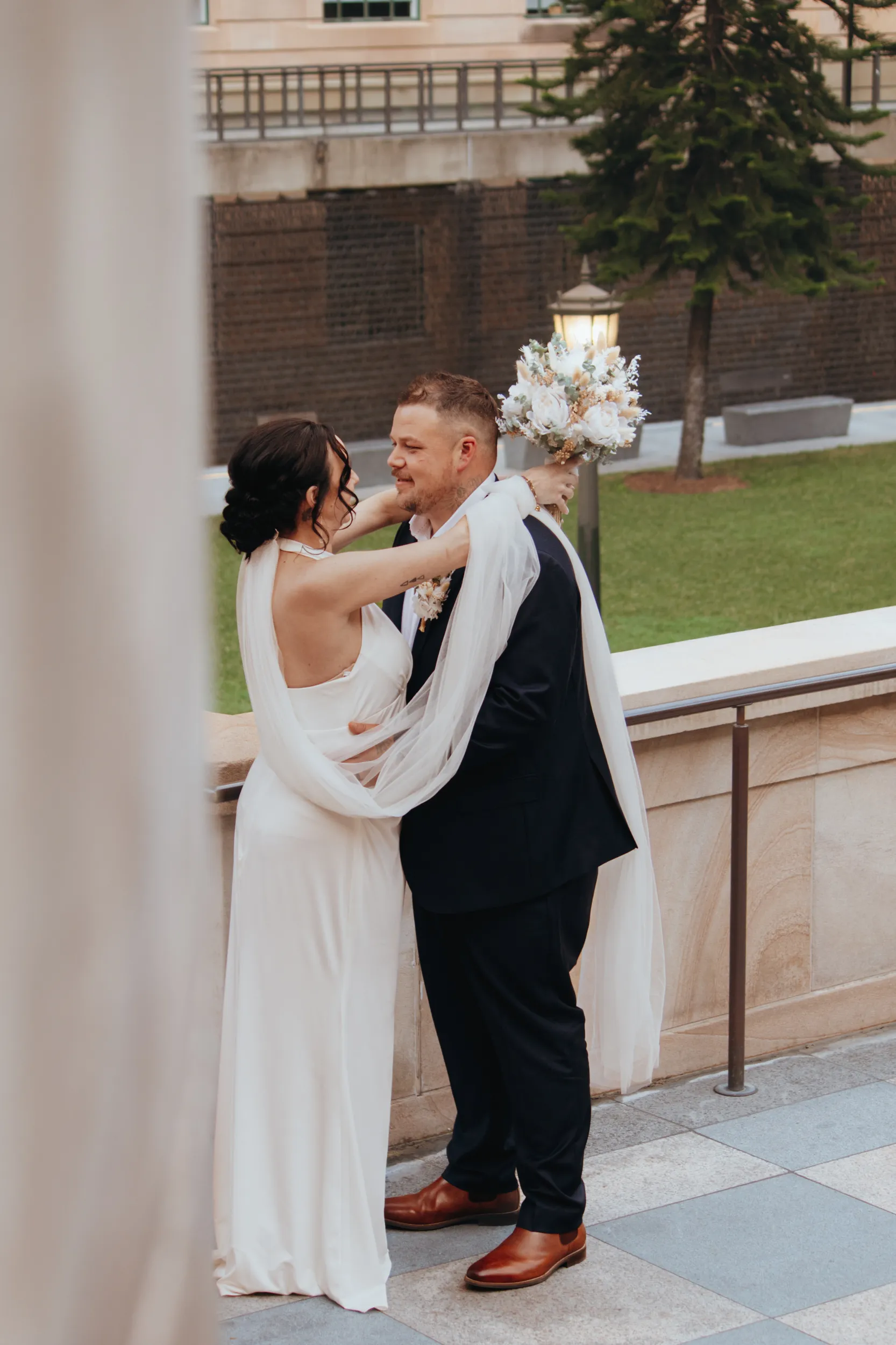 Tender wedding moment with bride in flowing white dress and veil embracing groom on elegant terrace