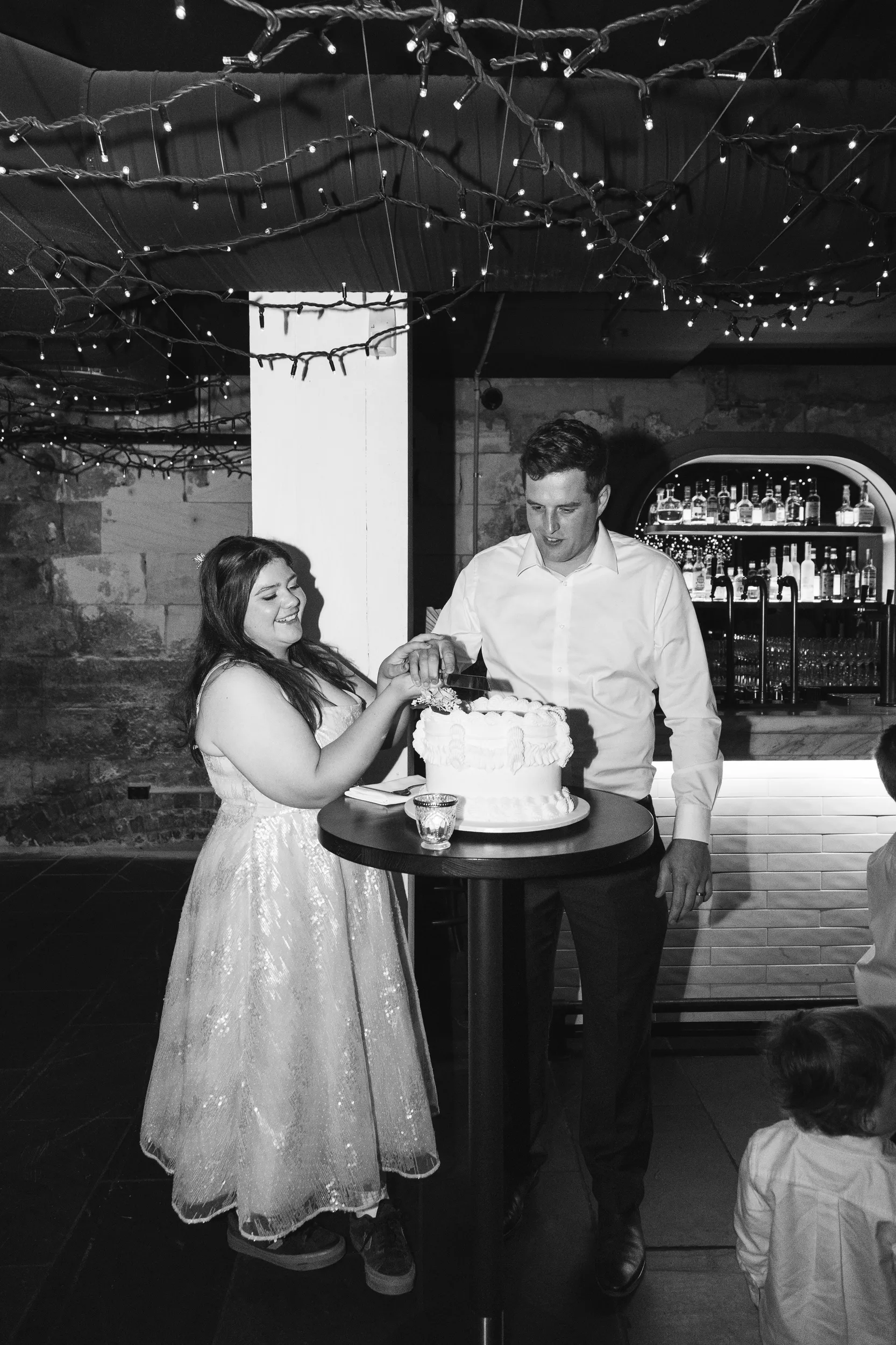 Black and white wedding cake cutting moment in rustic venue with string lights and exposed brick