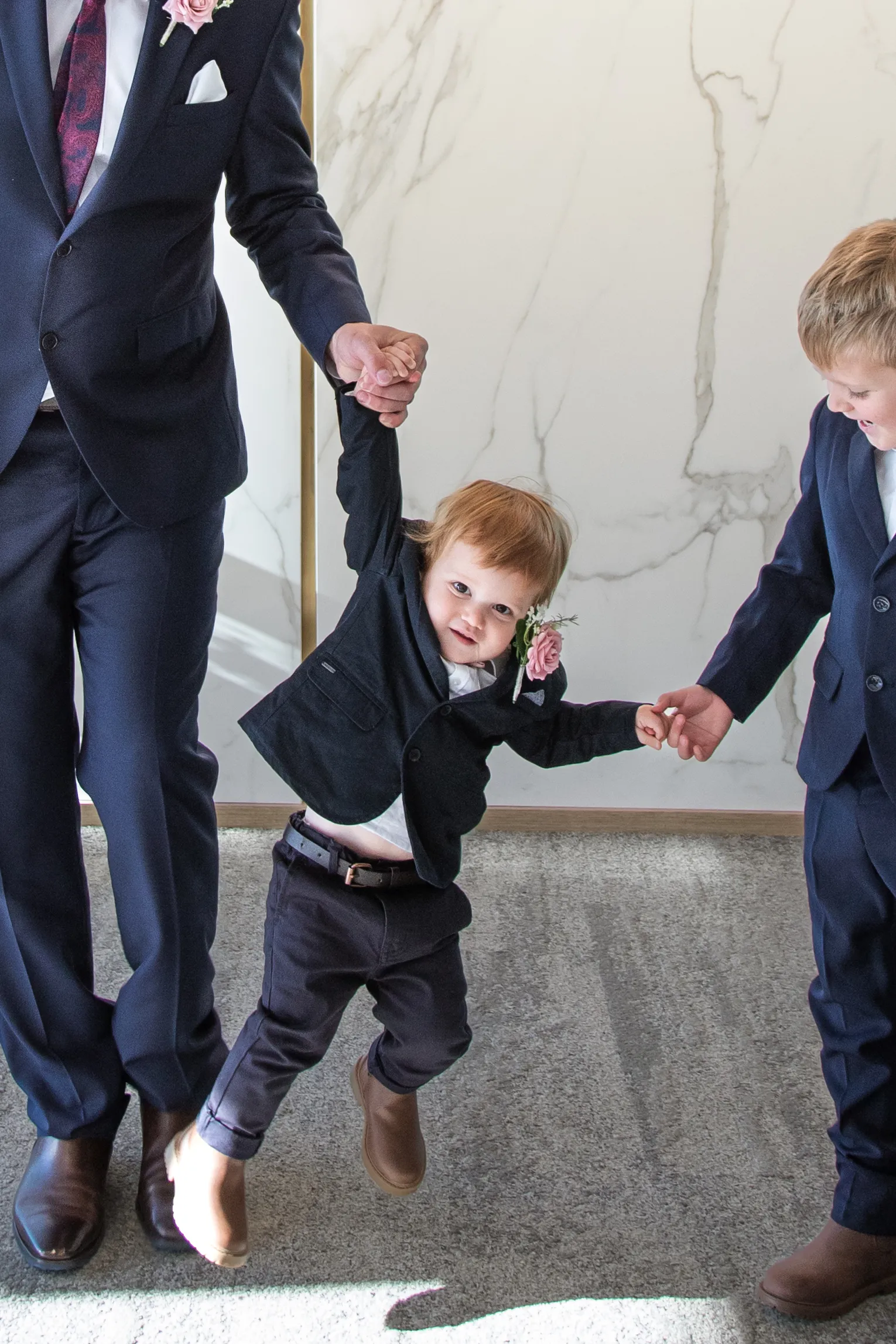 Heartwarming wedding moment with young child in navy suit and pink boutonniere being lifted by family members