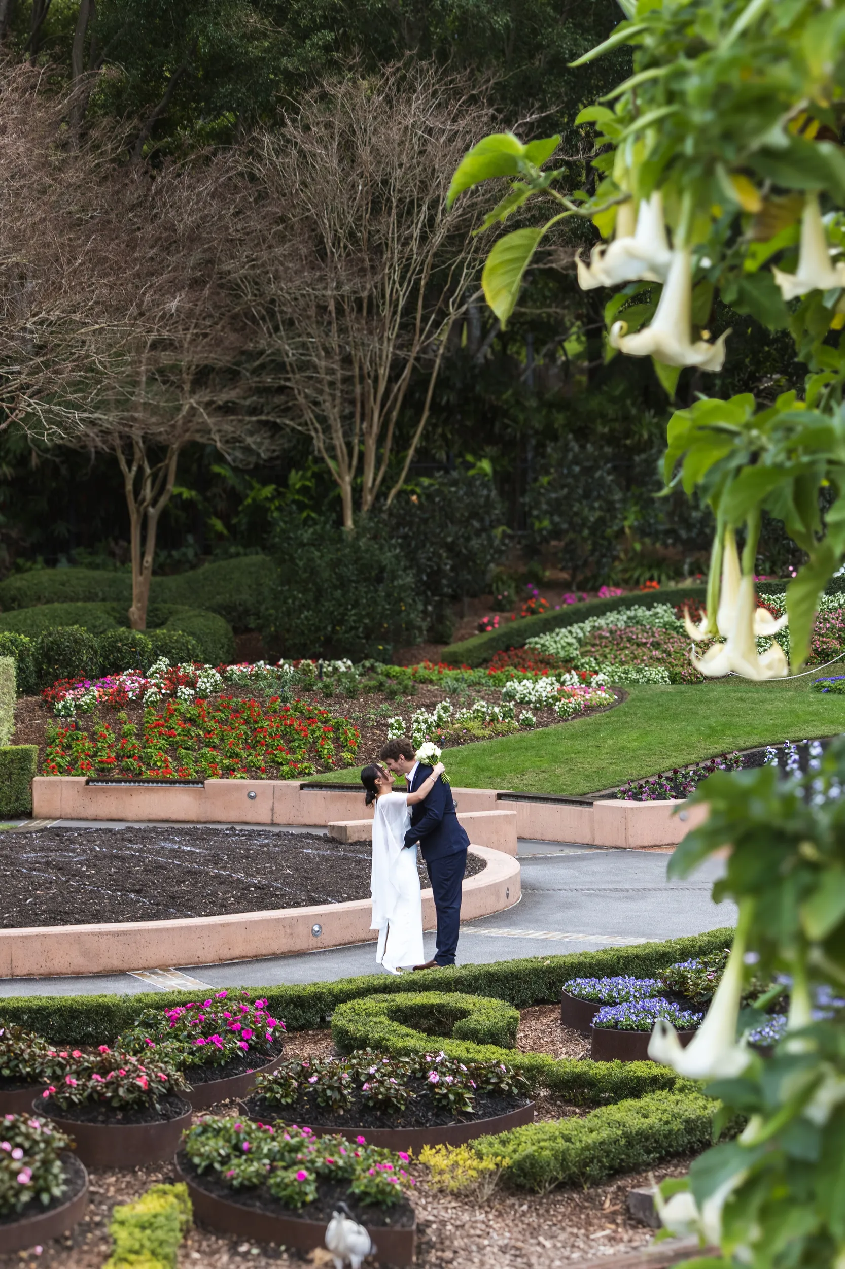 Beautiful wedding portrait of couple embracing in elaborate formal garden with colorful flower beds and tropical plants