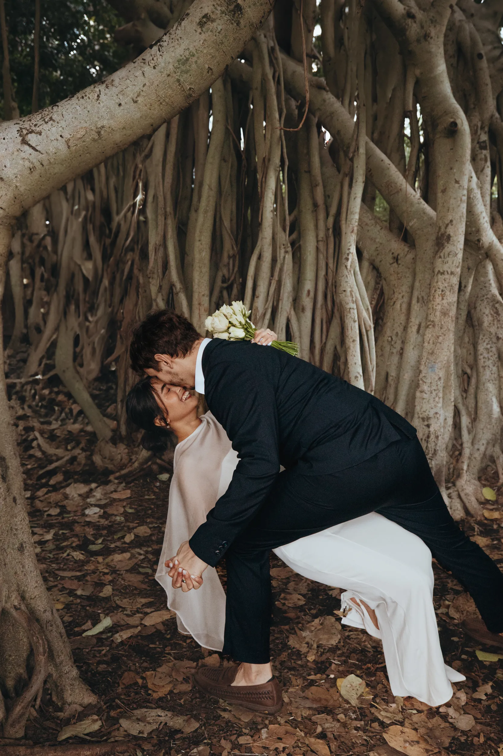 Playful wedding moment with couple laughing together under dramatic banyan tree aerial roots