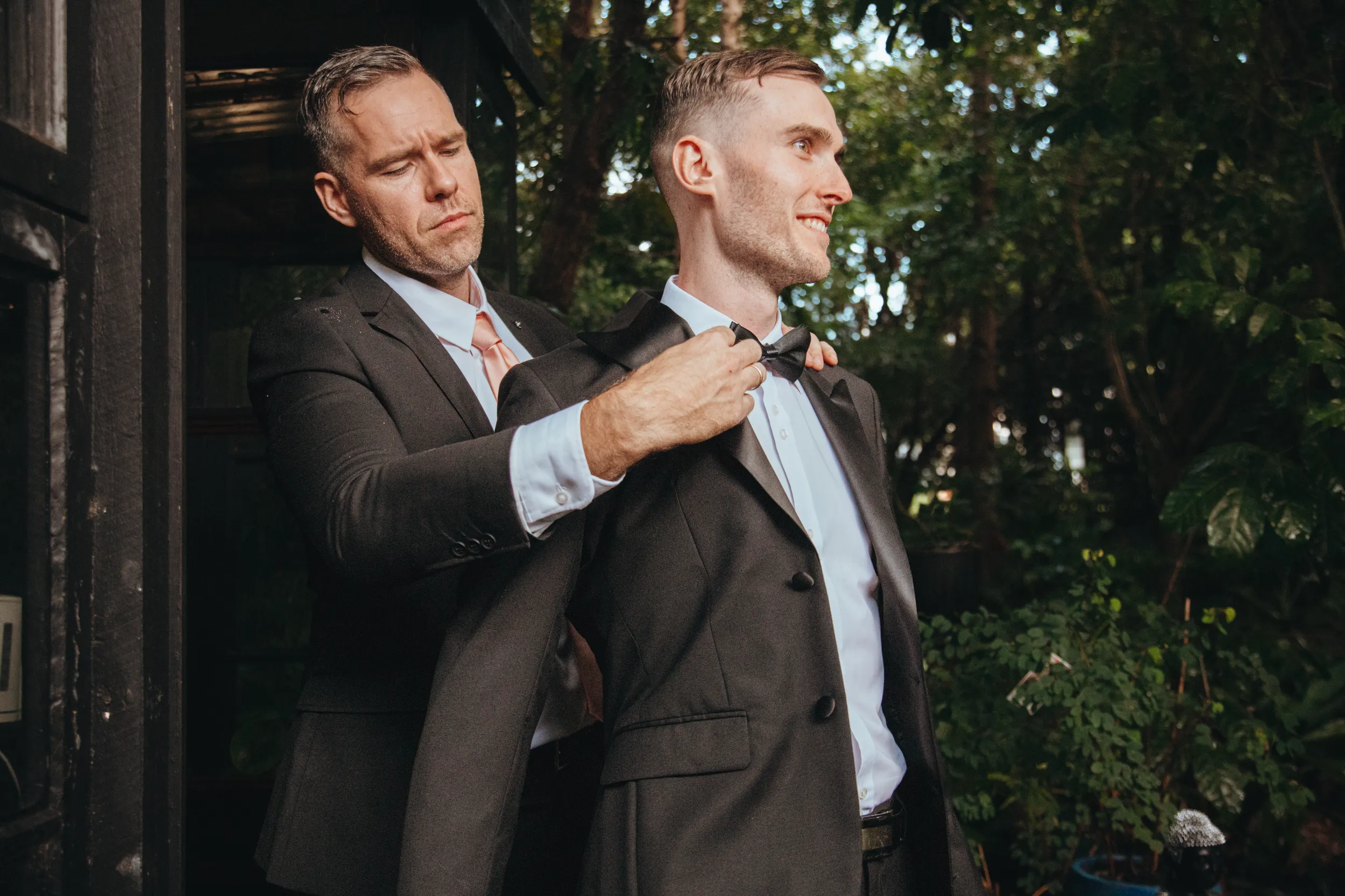 Touching wedding preparation moment with father helping groom adjust bow tie in outdoor garden setting